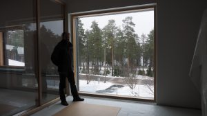 Tummana hahmona näkyvä mies ison ikkunan edessä, ikkunasta näkyy talvinen maisema.