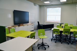 Luokkahuoneessa vihreitä pöytiä ja tuoleja, seinustalla iso näyttö, lattia on harmaa, sivuseinällä iso neliönmallinen ikkuna, joitakin mustia ja valkoisia kalusteita.