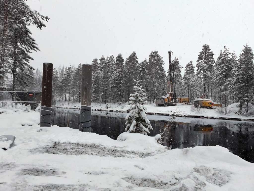Talvimaisemassa näkyy joki, jonka kuvaajanpuoleisella rannalla on kaksi pylvästä, toisella puolella jokea näkyy kaksi järeää työkonetta ja lumisia puita.