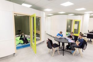 Kuvassa kaksi pyöreäkulmaista erillistilaa, joissa on keltainen lasiovi. Toisen ovi on auki ja siellä näkyy oppilas lukemassa. Erillistilan edessä pöytä, jossa kolme koululaista istuu ja opiskelee.