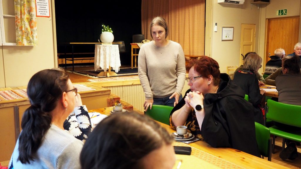 Naishenkilö seisoo pöydän äärellä, näkyvissä kolme pohdiskelevaa naista.